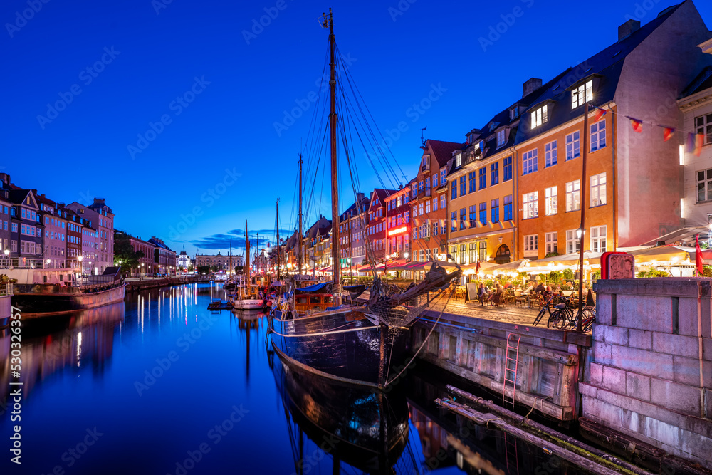 View of Nyhawn, the colorful houses next to the old port. Tourist visiting restaurants, cafes and ships in the canal at night. The most important sightseeing spot in Copenhagen, Denmark.