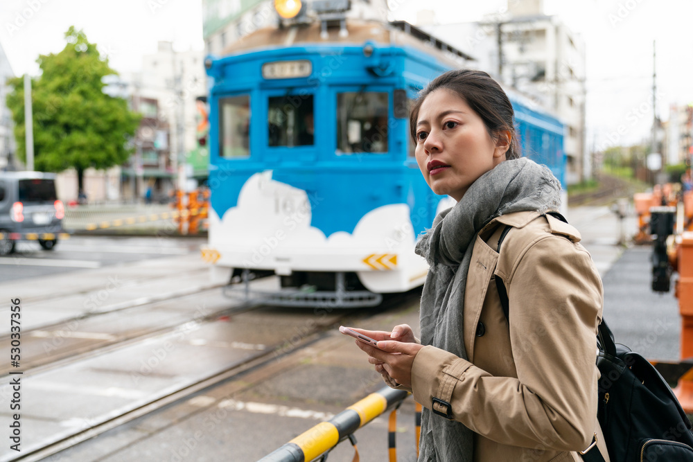female Asian Japanese traveler looking away into distance while standing by railway track barrier waiting for blue train to pass in Osaka city japan