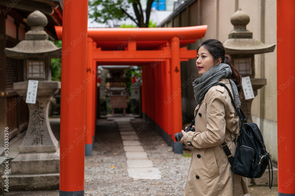 Asian Japanese girl photographer turning to look into distance at the entrance of rows of scarlet torii gates while traveling to Osaka Tenmangu Shrine in japan in spring
