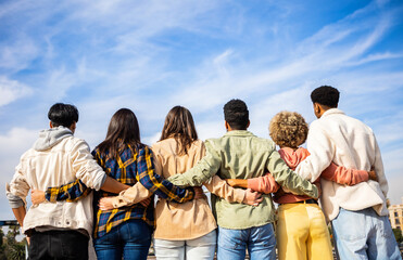 Rear view of young group of happy multiracial friends hugging each other standing in a row looking blue sky