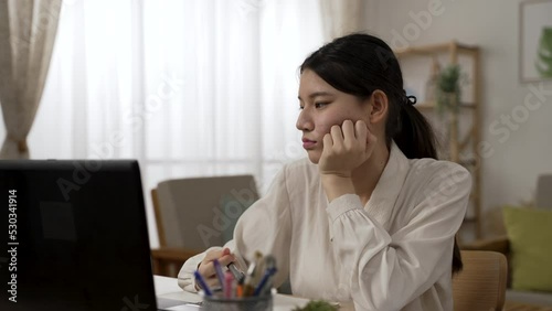 distracted asian woman employee is propping face and playing with pen while working from home, having no desire for work in the living room