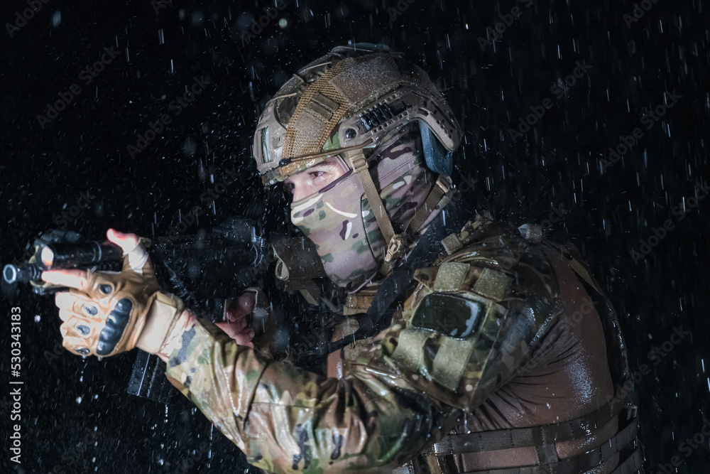 Fototapeta premium Army soldier in Combat Uniforms with an assault rifle, plate carrier and combat helmet going on a dangerous mission on a rainy night. 