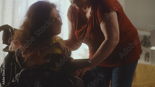 Aged woman caregiver helping woman with a spinal muscular atrophy setting a modern motorized wheelchair for comfortable sitting