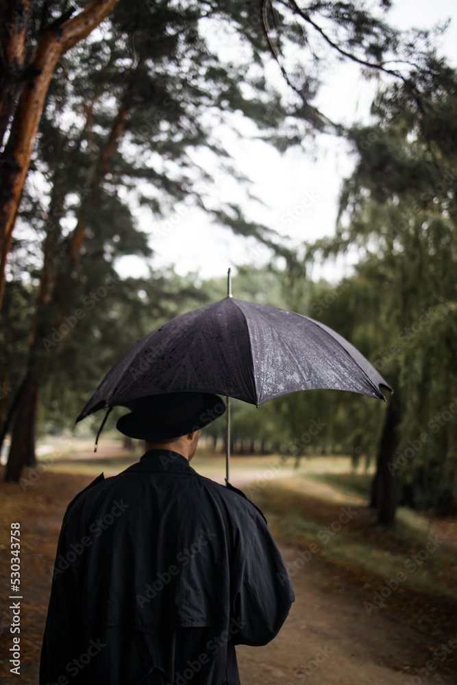 young english man in stylish vintage coat and black hat walking in rainy forest