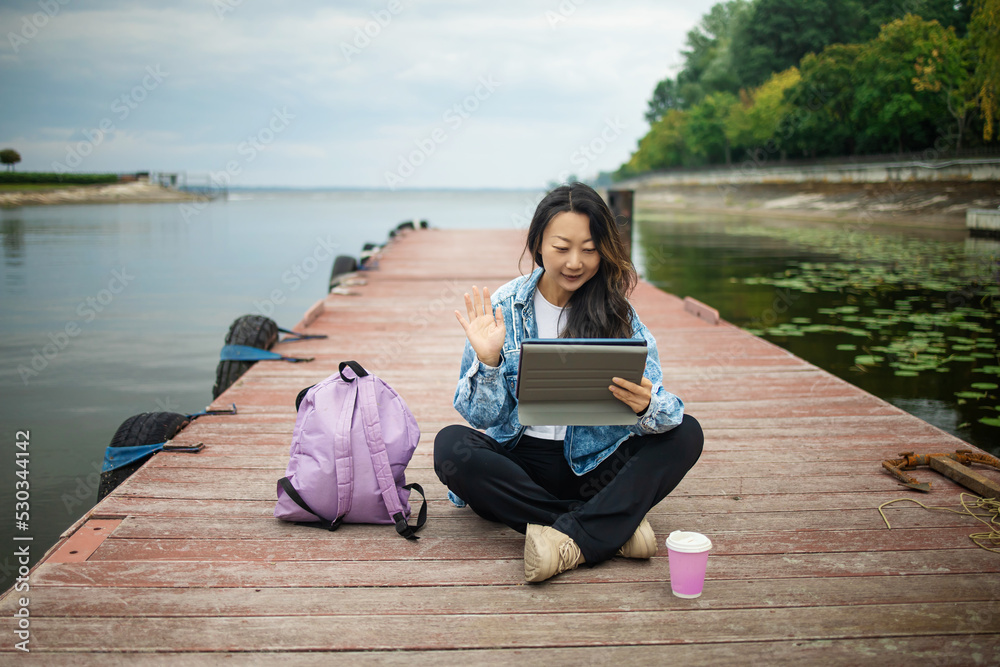 Stylish young beautiful asian woman traveler sits on the seashore and communicates by video call. 