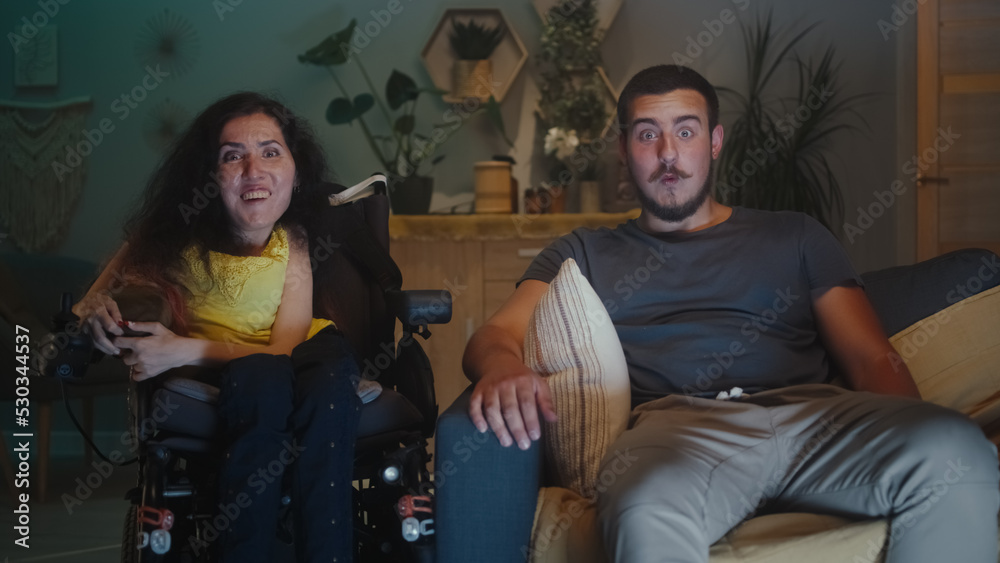 Shocked woman with physical disability in a wheelchair and man wondering and looking at each other, while watching TV show together at home in the evening