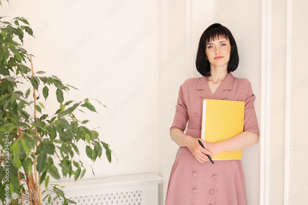 Attractive young woman in stylish soft pink dress is holding yellow folder of paperwork with mock up area looking in camera and standing in office with houseplants. Education, business, lifestyle Attractive young woman in stylish soft pink dress is holding yellow folder of paperwork with mock up area looking in camera and standing in office with houseplants. Education, business, lifestyle