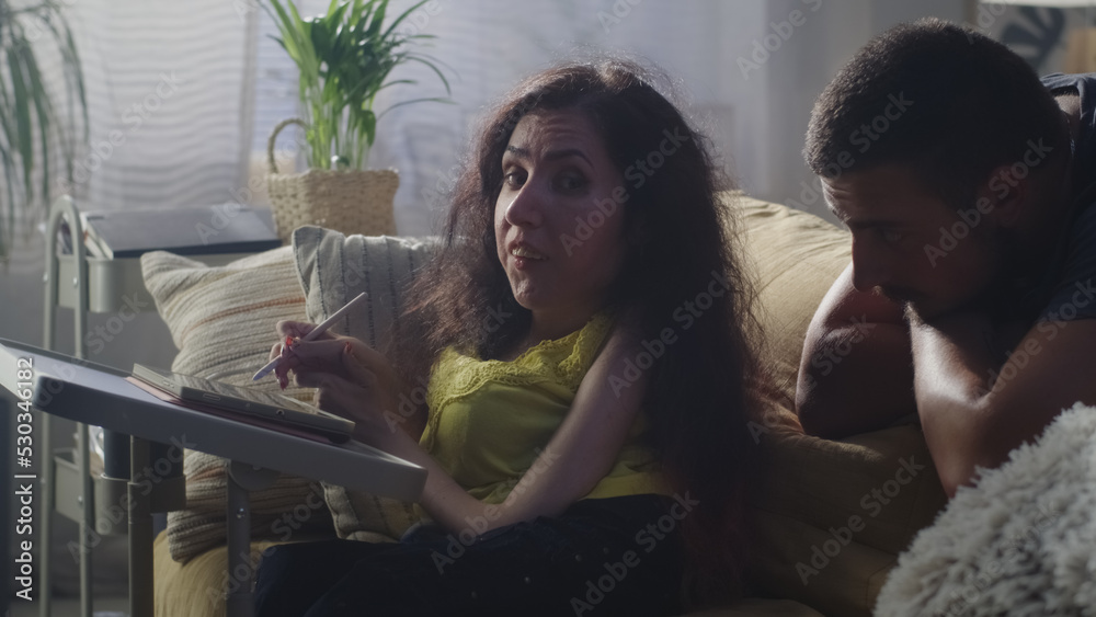 Woman with a disability drawing on digital tablet computer using pencil and talking with husband while sitting on a couch at the table at home Woman with a disability drawing on digital tablet computer using pencil and talking with husband while sitting on a couch at the table at home