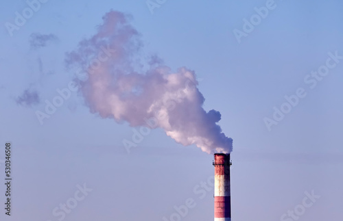 The plant's tall brick chimney emits a long cloud of thick white smoke against a blue sky.