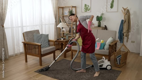 full length of a taiwanese woman in apron gloves is moaning in pain for she pulled her back muscles while cleaning the house with a hoover in the living room photo