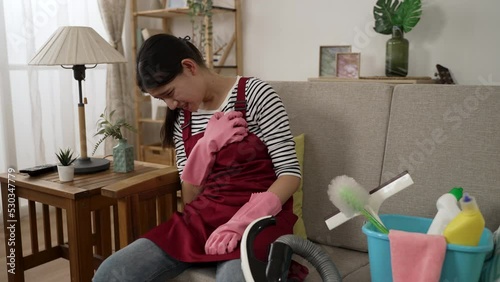asian cleaning woman wearing household apron is sitting on sofa with hand on chest and groaning in pain due to sharp pain from heart attack in the living room