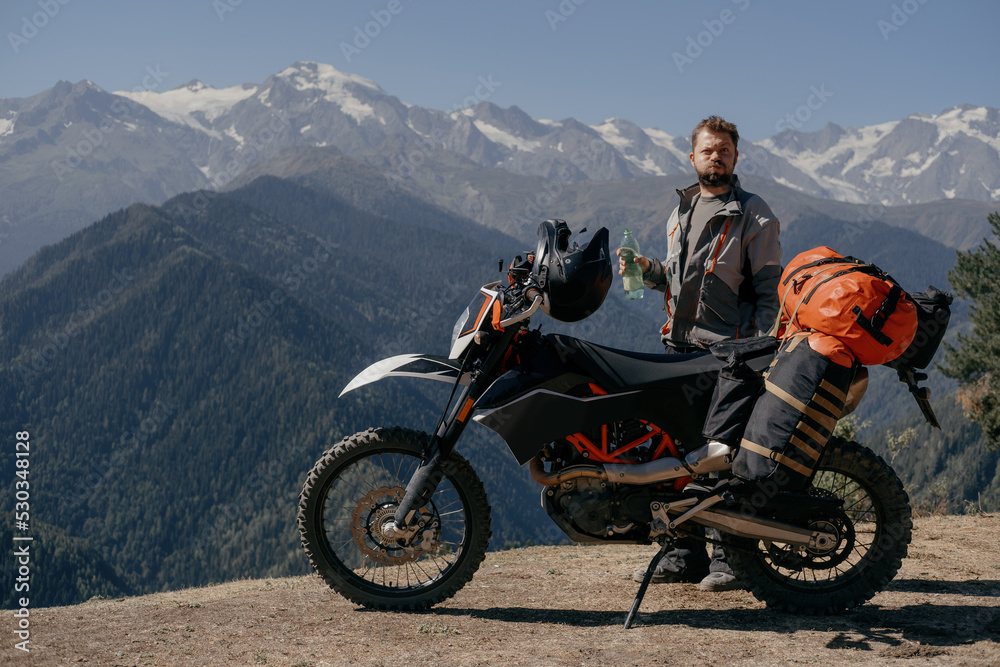 Biker man wearing beard and moto jacket standing near motorcycle and drinking water from bottle with funny face during sunny day in beautiful high mountains Biker man wearing beard and moto jacket standing near motorcycle and drinking water from bottle with funny face during sunny day in beautiful high mountains