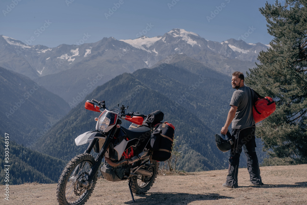 Fototapeta premium Male motorcyclist traveler standing in mountains near his motorcycle with large hermetic bag on his shoulder. Moto trip