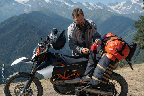 Man motorcyclist traveler standing next to dirt motorcycle and packing hiking bags with amazing mountains landscape on background