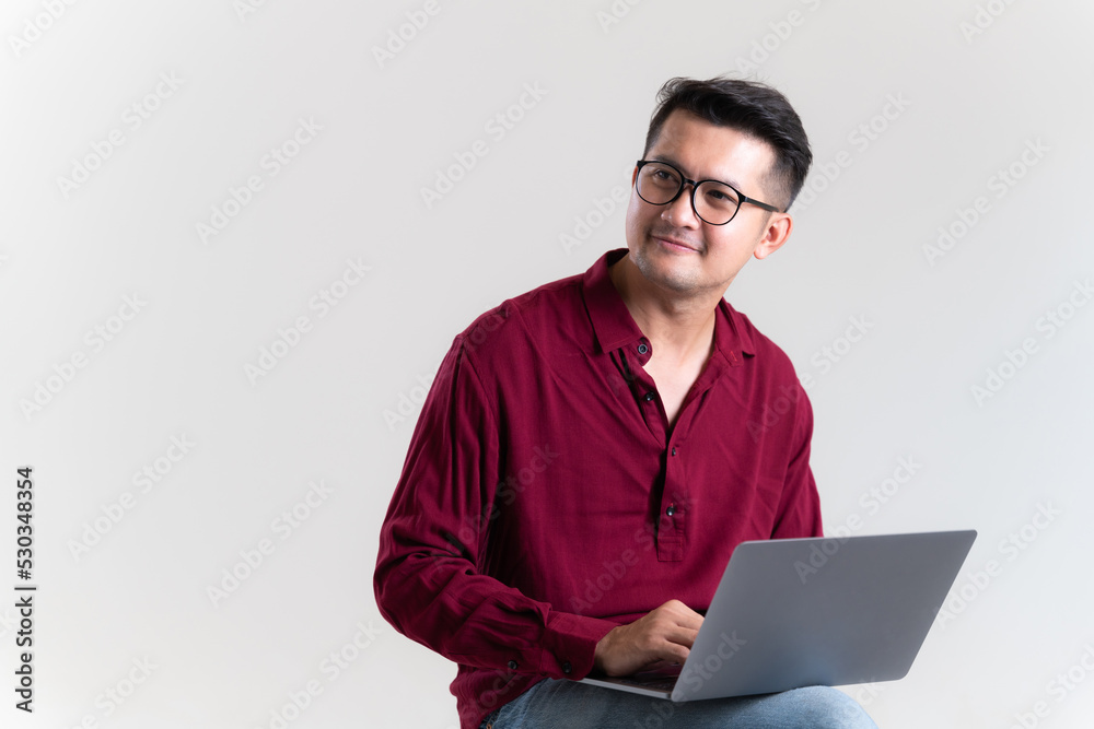 Asian man wearing a red shirt using laptop in studio grey background. The concept of freelance careers in the online world. Young Asian businessman looking to the future with a positive attitude. Asian man wearing a red shirt using laptop in studio grey background. The concept of freelance careers in the online world. Young Asian businessman looking to the future with a positive attitude.