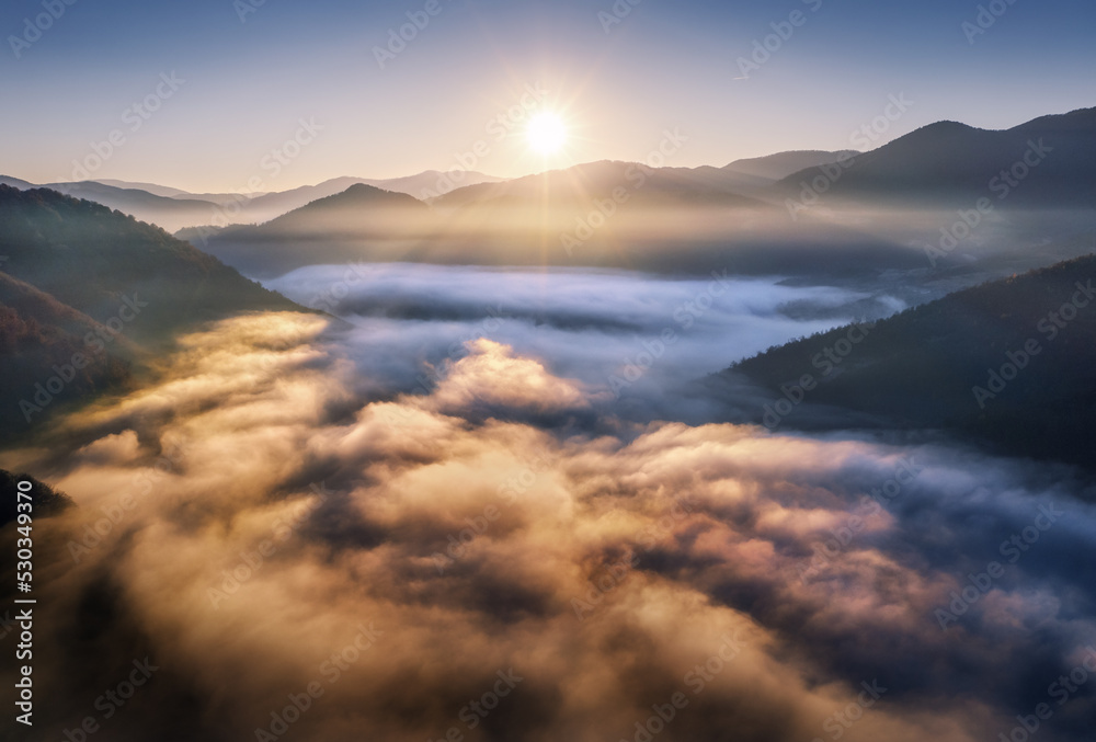 Mountains in low clouds at sunrise in autumn. Aerial view of mountain peak in fog in fall. Beautiful landscape with rocks, forest, sun, purple sky. Top view of mountain valley in clouds. Foggy hills