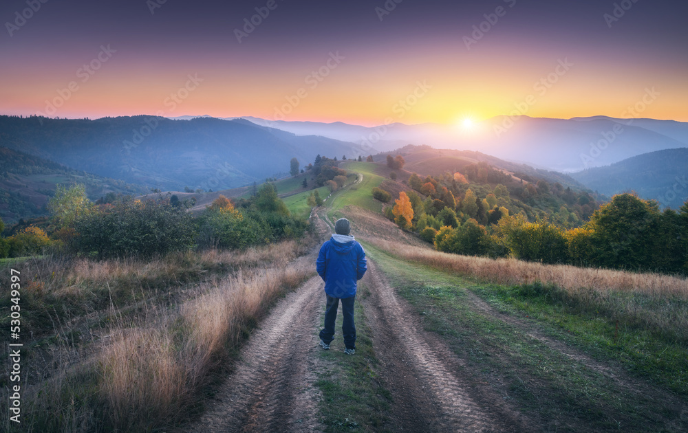 Man on the dirt road on the hill and mountains in fog at colorful sunset in autumn in Ukraine. Landscape with guy, foggy hills, green grass, meadows, trail, forest, beautiful sky in fall at dusk