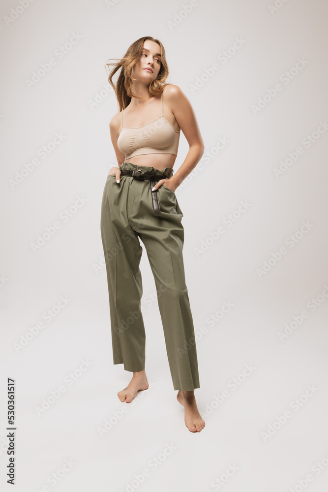 Fototapeta premium Beautiful woman with long curly hair and natural makeup wearing crop top and casual trousers posing over white background.