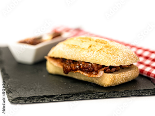 Crispy bacon in a sourdough roll with tomato relish on a slate with gingham napkin isolated against white background