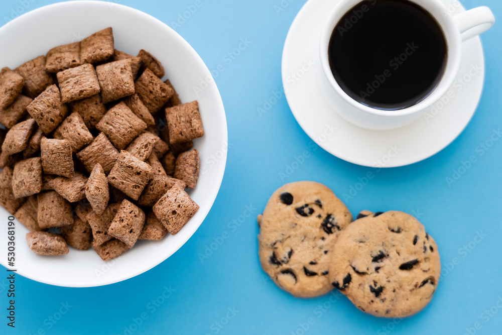 top view of cereal puffs in white bowl near cup of coffee and chocolate chip cookies isolated on blue. top view of cereal puffs in white bowl near cup of coffee and chocolate chip cookies isolated on blue.