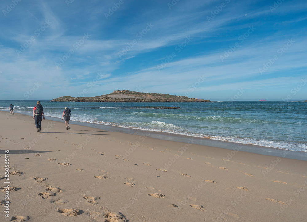 View of Praia da Ilha do Pessegueiro sand beach with group of hikers, ocean waves and Fort of Pessegueiro small island at Rota Vicentina coast near Porto Covo, Portugal. View of Praia da Ilha do Pessegueiro sand beach with group of hikers, ocean waves and Fort of Pessegueiro small island at Rota Vicentina coast near Porto Covo, Portugal.