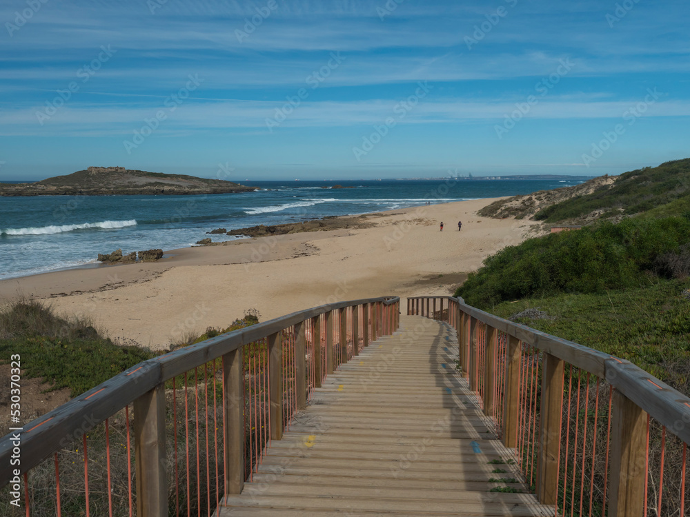 Obraz premium wooden bridge or sidewalk leading to Praia da Ilha do Pessegueiro sand beach with group of hikers, ocean waves and Fort of Pessegueiro small island at Rota Vicentina coast near Porto Covo, Portugal.
