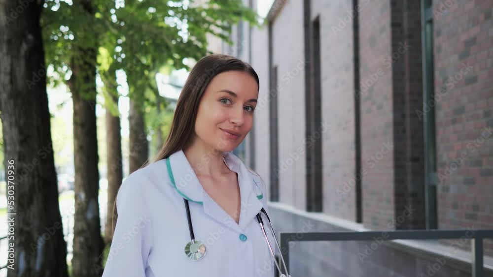 Beautiful long hair woman doctor wearing white medical coat, stethoscope turning looking at camera outdoor. Smiling female physician posing near hospital. Positive practitioner close up face portrait. Beautiful long hair woman doctor wearing white medical coat, stethoscope turning looking at camera outdoor. Smiling female physician posing near hospital. Positive practitioner close up face portrait.