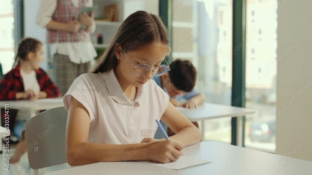 Exam in elementary school. Beautiful brunette asian scoolgirl wearing glasses sits at a first desk in the classroom and fills out tests. Education children. Exam in elementary school. Beautiful brunette asian scoolgirl wearing glasses sits at a first desk in the classroom and fills out tests. Education children.
