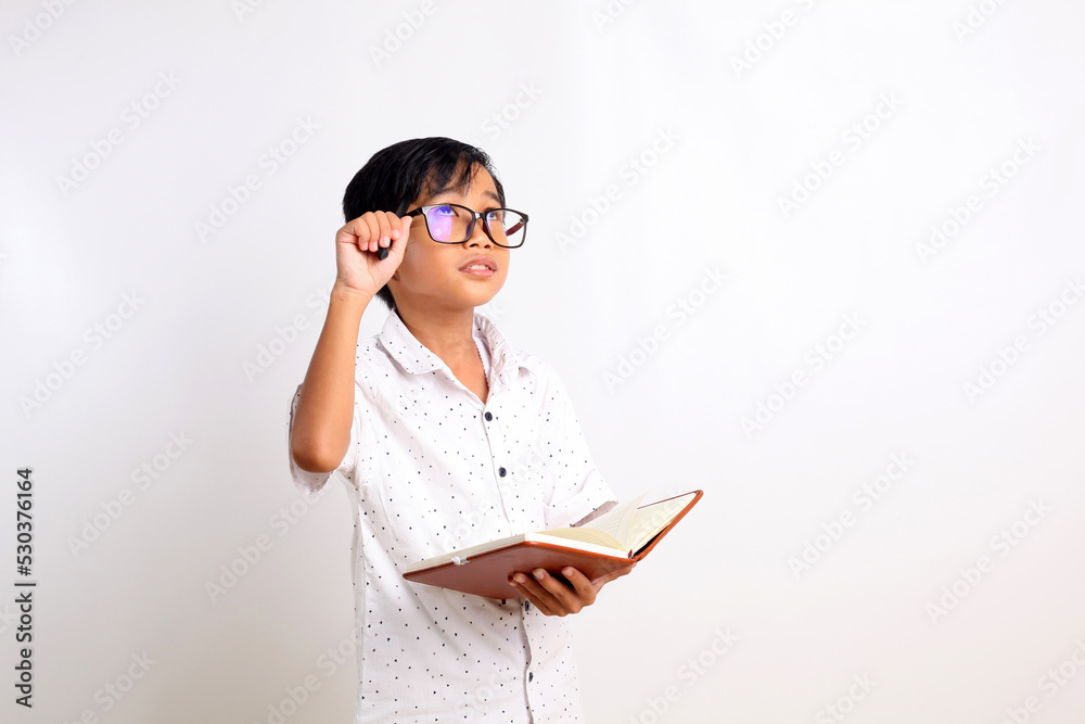 Smart asian schoolboy thinking while holding a book and looking up. Isolated on white background