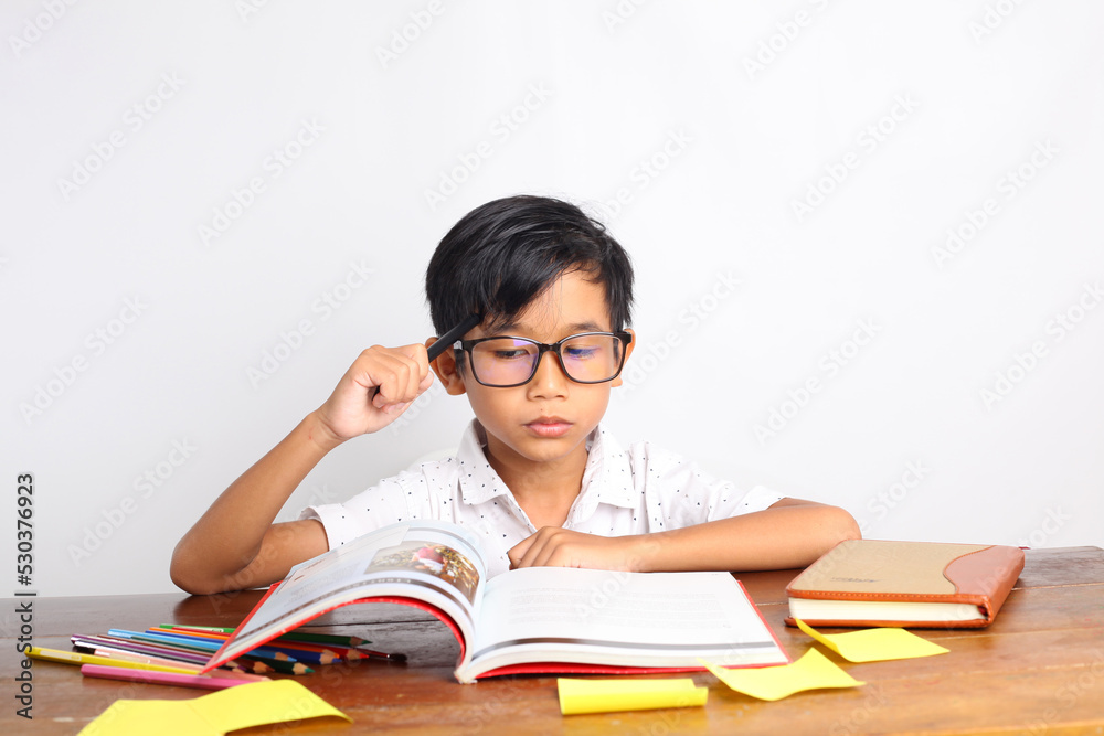 Thoughtful asian schoolboy studying in classroom while reading a book. Isolated on white background Thoughtful asian schoolboy studying in classroom while reading a book. Isolated on white background