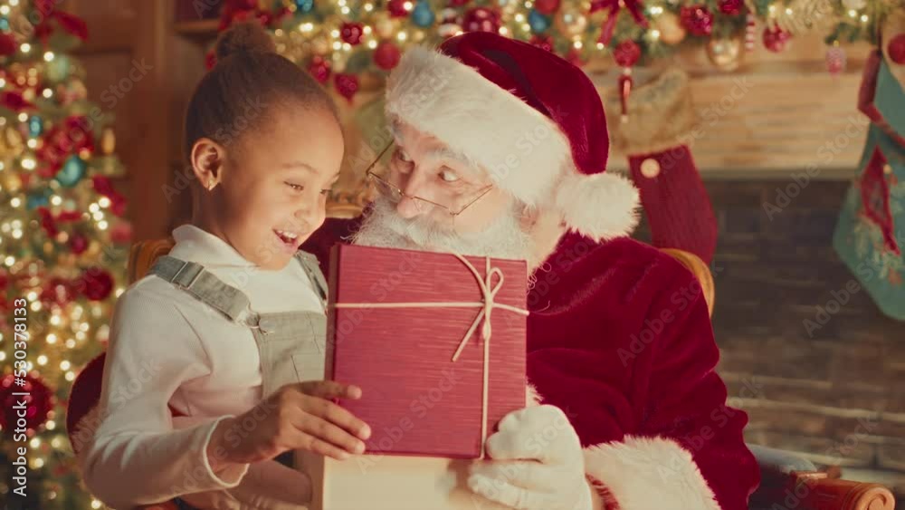 Medium slowmo of surprised Santa and pretty little African American girl opening sparkling Christmas present box together Medium slowmo of surprised Santa and pretty little African American girl opening sparkling Christmas present box together