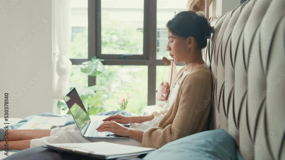 Happy young Asian girl with white cream cardigan on bed focus computer laptop full of paperwork messy document work idea in cozy bedroom at home in morning. Stay quarantine, Work from home concept. Happy young Asian girl with white cream cardigan on bed focus computer laptop full of paperwork messy document work idea in cozy bedroom at home in morning. Stay quarantine, Work from home concept.