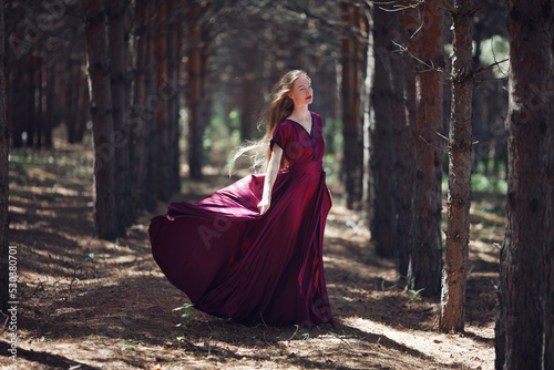 A tall, slender woman with long blond hair walks in the woods in a burgundy dress. In the pine forest.