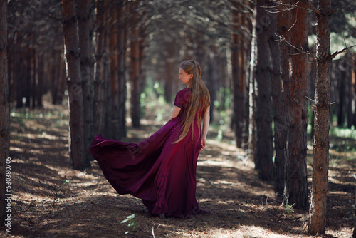 A tall, slender woman with long blond hair walks in the woods in a burgundy dress. In the pine forest.