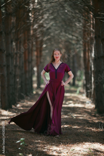 A tall, slender woman with long blond hair walks in the woods in a burgundy dress. In the pine forest.