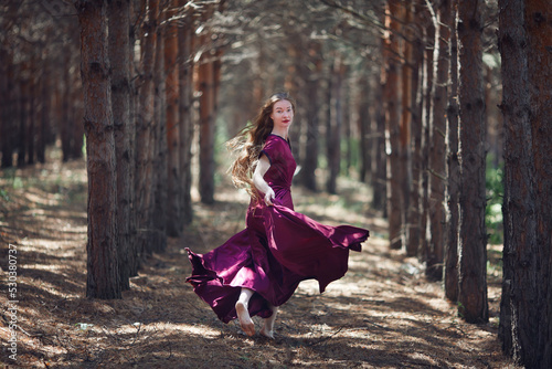 A tall, slender woman with long blond hair walks in the woods in a burgundy dress. In the pine forest.