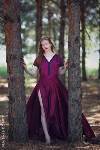 A tall, slender woman with long blond hair walks in the woods in a burgundy dress. In the pine forest.
