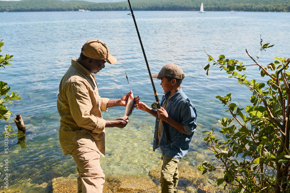 Side view of happy mature black man in activewear showing fish to his grandson with rod while both standing against waterside Side view of happy mature black man in activewear showing fish to his grandson with rod while both standing against waterside