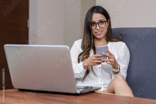 Latin woman typing on her cell phone while looking at the camera, working at home