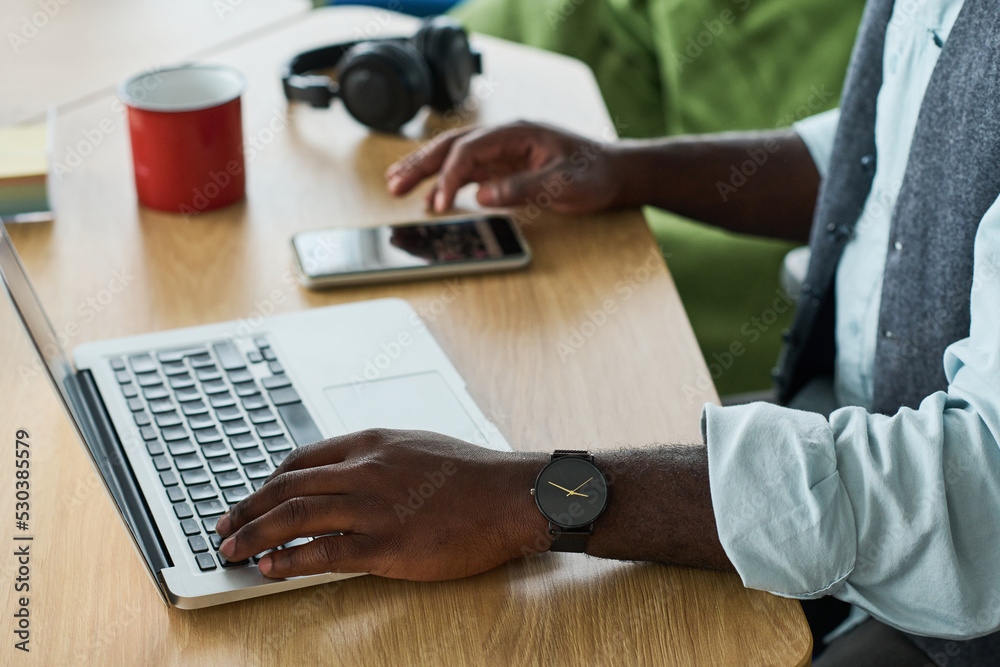 Hands of young black man in formalwear keeping hand over laptop keypad while scrolling in smartphone on workplace