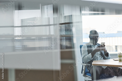 Young serious African American businessman in formalwear scrolling in smartphone while sitting by workplace in office with transparent walls