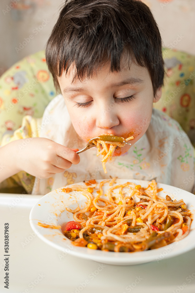 Child eating spaghetti with vegetables. Kid having fun eating. Brown haired boy with face covered in sauce. Weekend, warm and cozy scene in the kitchen Child eating spaghetti with vegetables. Kid having fun eating. Brown haired boy with face covered in sauce. Weekend, warm and cozy scene in the kitchen