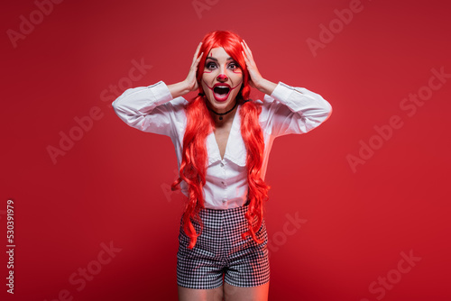 shocked woman in halloween makeup touching colored hair and screaming isolated on red.