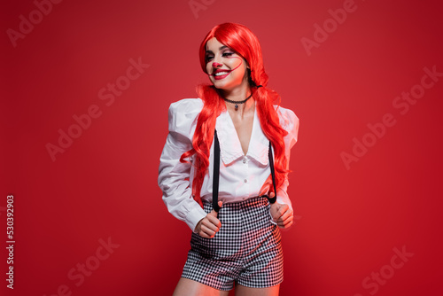 redhead woman with clown makeup posing in white blouse and shorts with suspenders isolated on red.