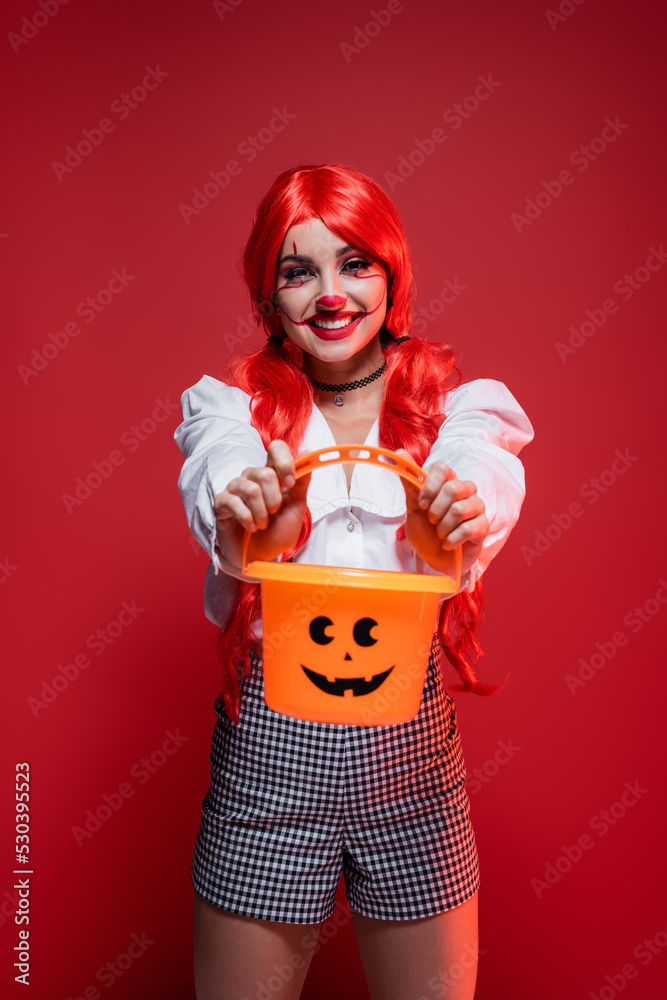 cheerful woman in clown makeup and bright wig showing halloween bucket isolated on red.