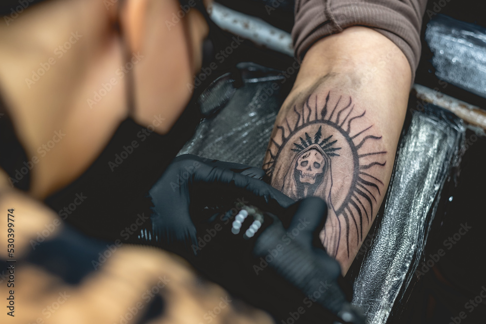 Detail of young tattoo artist girl with glasses and mask making a tattoo of 'La Santa Muerte' (Our Lady of Holy Death) in the arm of a woman with tattoo machine