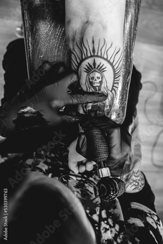 Detail of young tattoo artist girl with glasses and mask making a tattoo of 'La Santa Muerte' (Our Lady of Holy Death) in the arm of a woman with tattoo machine (in black and white)