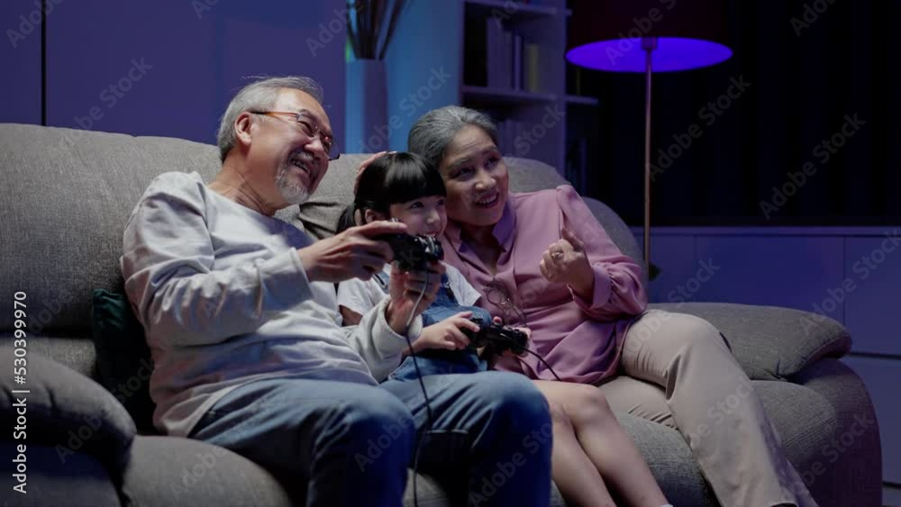Asian happy family play time. little girl with grandparents together playing video games use joystick control enjoy laughing fun in living room home at night. Happy child happiness lifestyle concept. Asian happy family play time. little girl with grandparents together playing video games use joystick control enjoy laughing fun in living room home at night. Happy child happiness lifestyle concept.