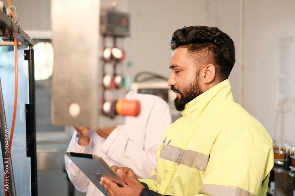 engineer worker working together with safety uniform and white helmet to work in industry factory handle tablet. Factory worker inspecting production line of drink production in factory. indian men engineer worker working together with safety uniform and white helmet to work in industry factory handle tablet. Factory worker inspecting production line of drink production in factory. indian men