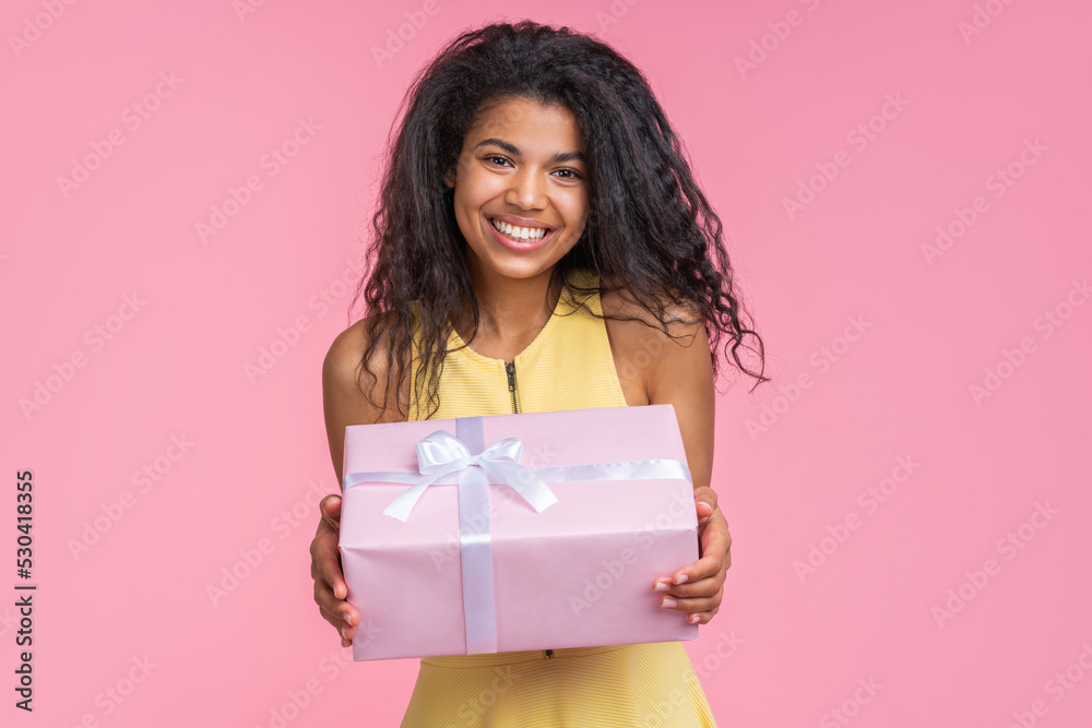 Studio portrait of happy smiling beautiful birthday girl posing with decorated present box in hands over pastel pink background Studio portrait of happy smiling beautiful birthday girl posing with decorated present box in hands over pastel pink background